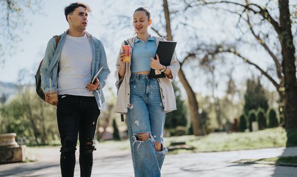 Zwei Oberstufenschüler arbeiten an einem Projekt, während sie im Freien in einem Campus-Umfeld spazieren gehen, chatten und Studienmaterialien in der Hand halten. - Foto, Bild