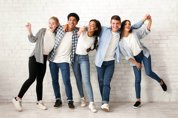 Skupina pěti multietnických teenagerů stojí v řadě u zdi z bílých cihel, usmívá se a směje. Jsou oblečeni v neformálním oblečení - Fotografie, Obrázek