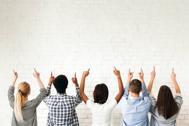 Um grupo de amigos está na frente de uma parede branca em branco, todos apontando para cima. Seus gestos sugerem que eles estão indicando ou chamando a atenção para algo acima deles, visão traseira - Foto, Imagem