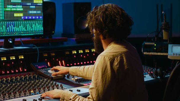 Music producer editing tracks with mixing console and audio software in studio, pressing key buttons and twisting knobs in control room. Sound engineer producing tunes and adjusting volume. Camera A. - Photo, Image