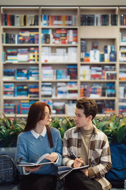 Two young adults, a male and a female, sit together in a well-stocked library, engaging in a collaborative study session. Their interaction suggests a mix of academic focus and friendly rapport. - Photo, Image