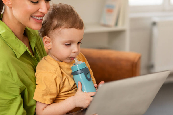 Una donna è seduta mentre tiene un bambino tra le braccia, multitasking utilizzando un computer portatile. Madre e figlio guardando cartone animato insieme, interno della casa, spazio copia - Foto, immagini
