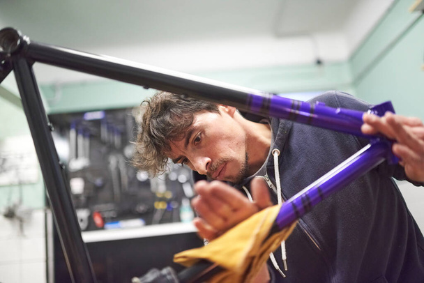 Spaanse jongeman die werkt aan een custom bike frame schilderij ontwerp in paars en zwart, een creatieve en technische handgemaakte proces, in zijn werkplaats. Echte mensen aan het werk. - Foto, afbeelding