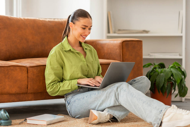 Nainen istuu lattialla, keskittyen kannettavan tietokoneen käyttöön hänen edessään. Hän esiintyy työssään, vakava ilme kasvoillaan. - Valokuva, kuva