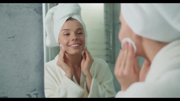 De vrouw met een handdoek om haar hoofd gebruikt haar vinger om haar gezicht voor de spiegel schoon te maken, zich concentrerend op elke wimper. - Video