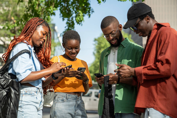 Inmerso por los teléfonos inteligentes grupo de jóvenes afroamericanos estudiantes miran fijamente en las pantallas de navegación en las redes sociales sin interactuar entre sí al aire libre. Amigos interesados con adicción al teléfono. - Foto, Imagen