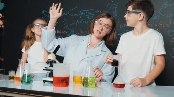 Blanke jongen mengen chemische vloeistof, terwijl de leraar geeft advies. Professionele instructeur dragen lab pak op zoek naar diverse student aan tafel met beker gevuld met gekleurde oplossing. Erutie. - Video