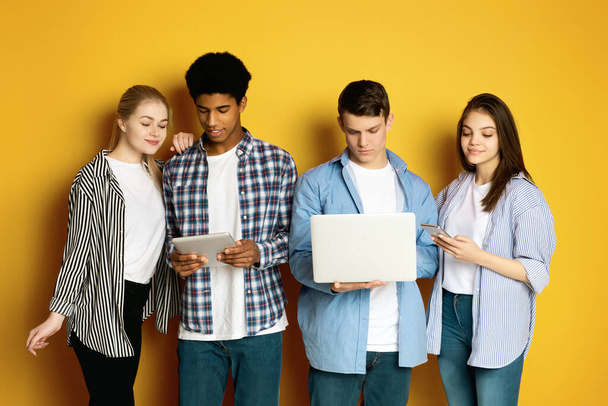 Quatro adolescentes multiétnicos enfrentam um cenário amarelo brilhante, cada um profundamente focado em sua própria peça de tecnologia.. - Foto, Imagem