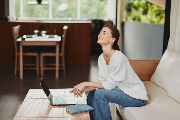Молодая женщина комфортно сидит на современном диване в своей уютной гостиной, используя свой ноутбук с улыбкой Окруженная современным интерьером, она наслаждается своими выходными, общаясь в Интернете, покупки, и - Фото, изображение