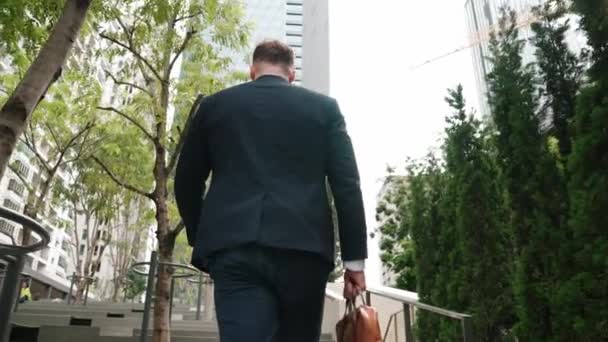 Rückenansicht eines kaukasischen Managers im formellen Anzug, der die Treppe hinaufgeht, während er seinen Koffer in der grünen Stadt hält. Professioneller Geschäftsmann, der zur Arbeit geht, während er die Treppe in der Öko-Stadt erklimmt. Urbane Pflanzen.. - Filmmaterial, Video