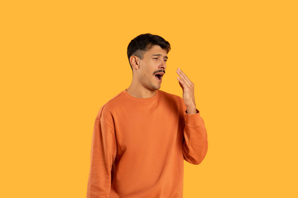 Un joven de cabello oscuro se levanta contra un fondo amarillo vivo, cubriéndose la boca con la mano mientras bosteza ampliamente, mostrando signos de fatiga o aburrimiento usando una sudadera naranja casual.. - Foto, imagen