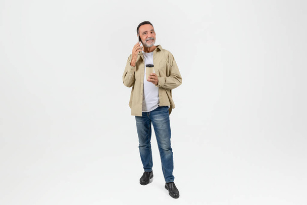 Un uomo anziano si alza casualmente e beve da una tazza termica, isolato su uno sfondo bianco, godendo di un momento di relax, parlando al telefono - Foto, immagini