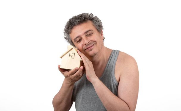 Un uomo di mezza età dai capelli grigi, arruffato e non rasato con una maglietta senza maniche tiene in mano una casa in legno in miniatura - Foto, immagini