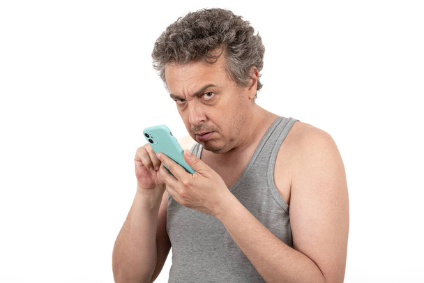 Um homem de meia-idade grisalho, desgrenhado e sem barba com uma camiseta sem mangas tem um smartphone - Foto, Imagem