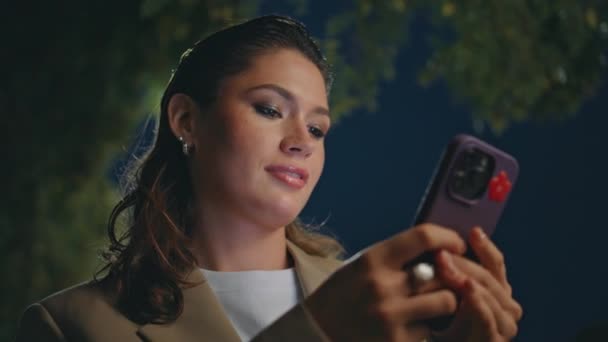 Ελκυστική κυρία περιήγηση εφαρμογή smartphone στέκεται κάτω από τα φώτα του δρόμου και μόνο από κοντά. Μια χαμογελαστή γυναίκα στέλνει μηνύματα στο κινητό της τη νύχτα. Πανέμορφη επιχειρηματίας που κανονίζει συνάντηση με κινητό τηλέφωνο. - Πλάνα, βίντεο