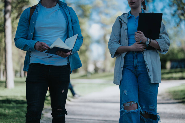 Twee vrienden, een mannelijke en een vrouwelijke student, wandelen buiten met academische essentials in een serene park omgeving. - Foto, afbeelding