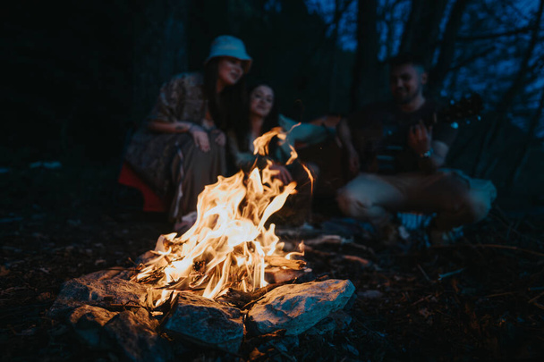 Tre amici si riuniscono intorno a un fuoco di campo ardente nella foresta, condividendo storie e suonando la chitarra sotto il cielo della sera. - Foto, immagini