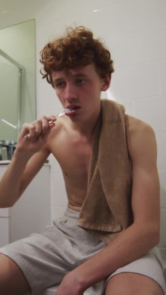 Вертикальний знімок молодого чоловіка, який сидить на туалеті у ванній кімнаті і чистить зуби під час ранкового похмілля вдома - Кадри, відео