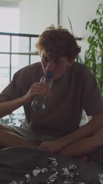 Вертикальний знімок молодого чоловіка з брудним волоссям, що сидить на незробленому ліжку з мішурою на ньому, п'є воду і дивиться на камеру під час ранкового похмілля - Кадри, відео