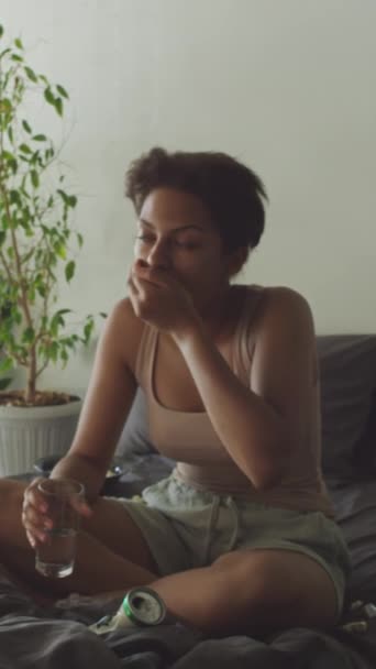 Κάθετη λήψη του άρρωστου κοριτσιού που πίνει χάπια και υποφέρει από ναυτία ενώ κάθεται στο κρεβάτι κατά τη διάρκεια του πρωινού στο σπίτι - Πλάνα, βίντεο