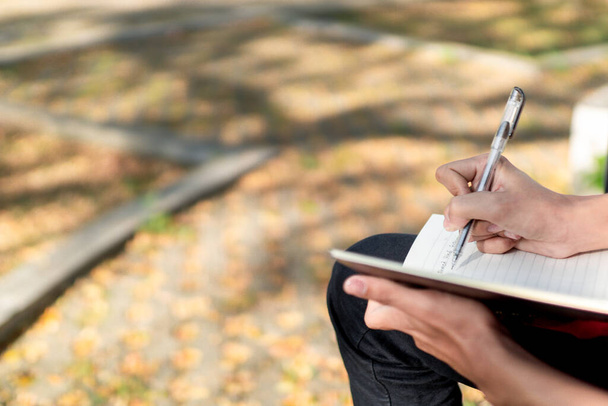 Νέος Ασιάτης σπουδάζει. Ένας άντρας γράφει σε ένα σημειωματάριο στο δημόσιο πάρκο. φοιτητής που κάθεται σε εξωτερικούς χώρους στο πάρκο της φύσης γράφοντας σημειώσεις στο σημειωματάριο. Άνδρας χέρι με στυλό γραφής το πρωί - Φωτογραφία, εικόνα