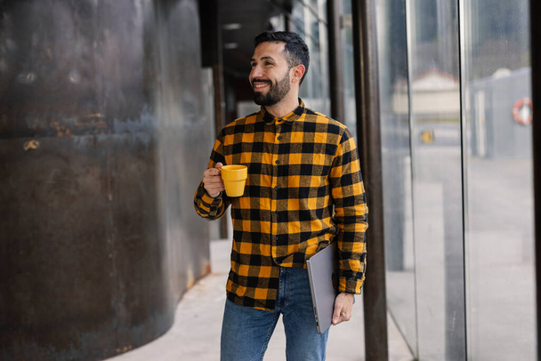 Homem hispânico alegre em seus 30 anos, segurando uma xícara de café e tablet, desfruta de um momento em uma calçada da cidade. - Foto, Imagem