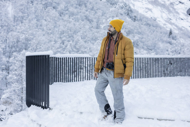 Ένας γενειοφόρος άντρας με κίτρινο σκουφί και μονωμένο σακάκι περπατά μέσα από βαθύ χιόνι, με κάμερα στο χέρι, σε ένα εκπληκτικό χιονισμένο ορεινό σκηνικό. - Φωτογραφία, εικόνα