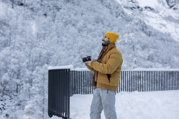 Vreugdevolle fotograaf in een gele muts en warme jas legt met zijn camera de schoonheid van een besneeuwd berglandschap vast. - Foto, afbeelding