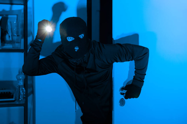 Una imagen que muestra a un ladrón usando un pasamontañas y usando una linterna para hurgar en un estante de un apartamento durante la noche - Foto, imagen