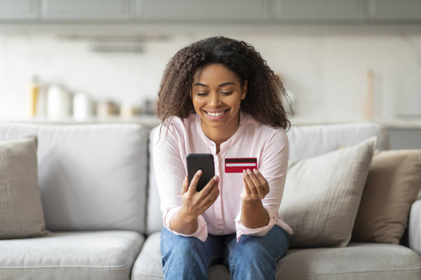 Cheerful black lady sitting at home, using a credit card and smartphone, signifying online shopping and payment - Photo, Image