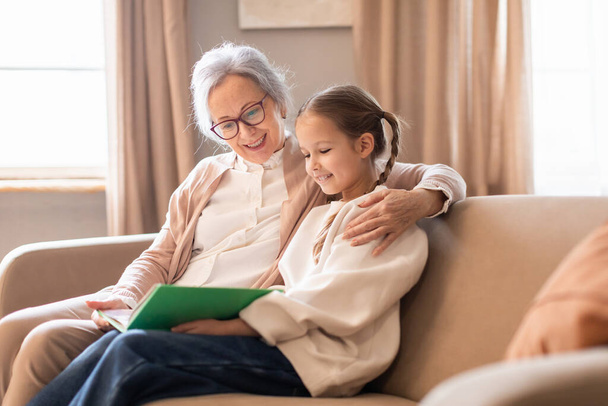Starsza kobieta i młoda dziewczyna siedzą na kanapie w domu, uczestnicząc w rozmowie podczas czytania książki razem - Zdjęcie, obraz