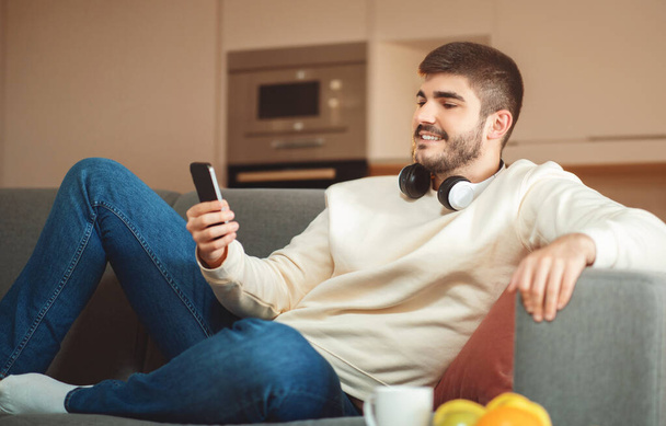 Расслабленный мужчина сидит на диване, держа сотовый телефон руками. Он сфокусирован на экране, взаимодействуя с устройством. - Фото, изображение