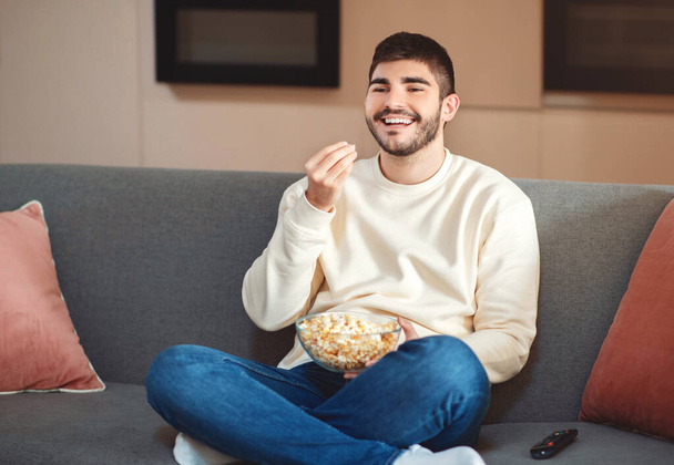 Un hombre está sentado cómodamente en un sofá, comiendo un tazón de palomitas de maíz. Parece relajado mientras disfruta de su merienda en el acogedor entorno. - Foto, imagen