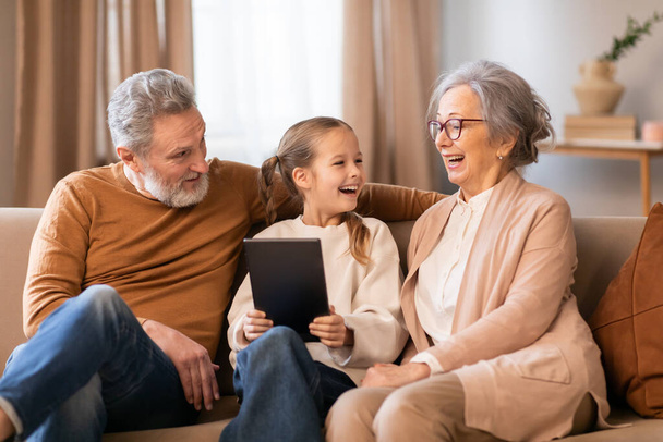 Mladá dívka, sedící mezi svými prarodiči, s nimi prožívá radostný okamžik na pohodlném gauči, když spolu doma používají digitální tablet - Fotografie, Obrázek