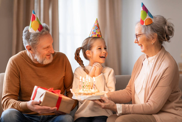 Μια νεαρή κοπέλα, φορώντας ένα εορταστικό καπέλο κόμμα, λαμβάνει χαρούμενα μια τούρτα γενεθλίων από τη γιαγιά της, ενώ μια εξίσου χαρούμενη παππούς κοιτάζει για κρατώντας ένα δώρο - Φωτογραφία, εικόνα
