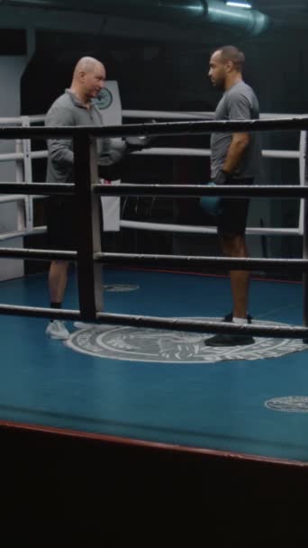 Afroamerykanin w rękawiczkach bokserskich stoi na ringu i rozmawia z trenerem. Kaukaski trener wyjaśnia techniki walki i skonsultować bokser przed konkurencją. Strzał pionowy - Materiał filmowy, wideo