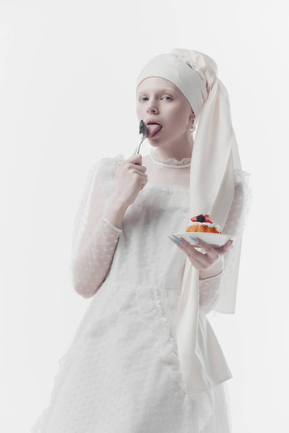 Woman wearing white embroidered dress and a stylish turban, as medieval person eating sweet dessert against white studio background. Concept of history, renaissance art, comparison of eras, vintage. - Photo, Image