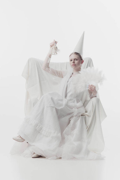 Vrouw in wit kostuum met conische hoed, ziet eruit als middeleeuwse persoon, met veren ventilator en rinkelen op kleine bel tegen witte studio achtergrond. Concept van renaissance kunst, vergelijking van tijdperken. Steun - Foto, afbeelding