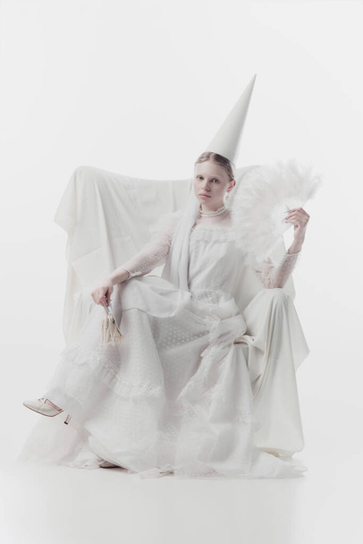 Femme en costume blanc avec chapeau conique, ressemble à une personne médiévale, tenant ventilateur à plumes et petite cloche ornée sur fond de studio blanc. Concept d'histoire, art de la renaissance, comparaison des époques. - Photo, image