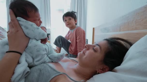 Moeder houdt pasgeboren baby zoon terwijl ze in bed ligt in de ochtend als 5-jarige jongen broer zitten naast haar, authentieke levensstijl familie scene - Video