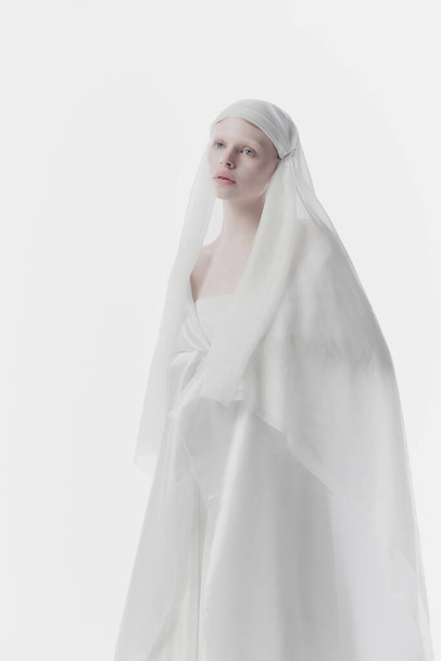 Young, calm woman shrouded in gossamer white fabric, creating ghostly yet angelic appearance against white studio background. Concept of history, renaissance art, comparison of eras, vintage. - Photo, Image