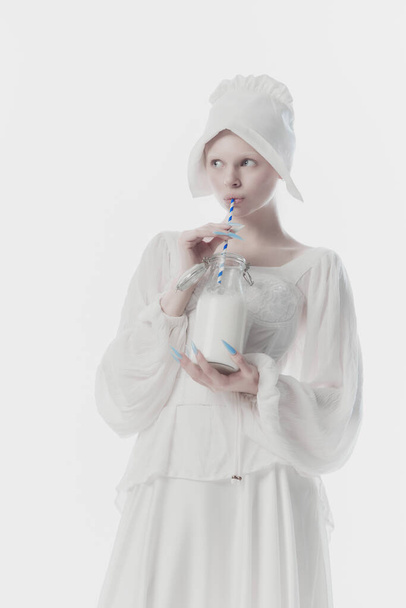 Gyönyörű fiatal nő öltözött középkori személy, szobalány fehér történelmi ruha iszik tej ellen fehér stúdió háttér. Történelmi fogalom, reneszánsz művészet, korok összehasonlítása, évjárat. - Fotó, kép
