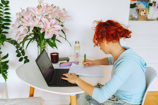portrait red-haired, curly teenage girl, with headband on hair, sitting at round white table in room, using computer, studying, writing notes, on table next to there is bouquet of lilies in vase - Photo, Image