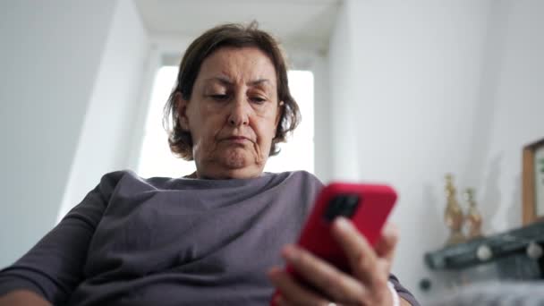 Mujer mayor sosteniendo el dispositivo de teléfono celular leyendo contenido en línea mientras está sentada en el sofá en la sala de estar, expresión seria de la persona de 70 años mirando la pantalla - Imágenes, Vídeo