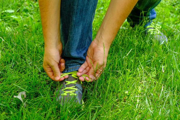 Mies sitomassa kengännauhojaan lenkkareihinsa vihreää ruohoa.
 - Valokuva, kuva