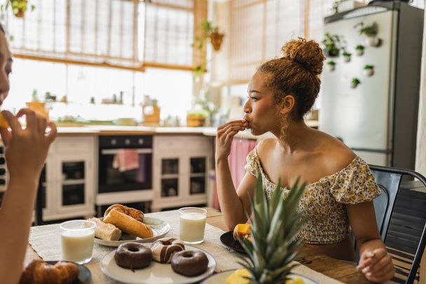 Egy fiatal vegyes fajú nő élvezi a finom süteményeket a reggeli beszélgetés során egy barátjával egy melegen megvilágított konyhában, tele friss gyümölcsökkel és péksüteményekkel.. - Fotó, kép
