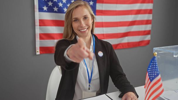 Mujer sonriente apunta a cámara en un centro de votación con bandera americana, mostrando participación cívica. - Foto, imagen