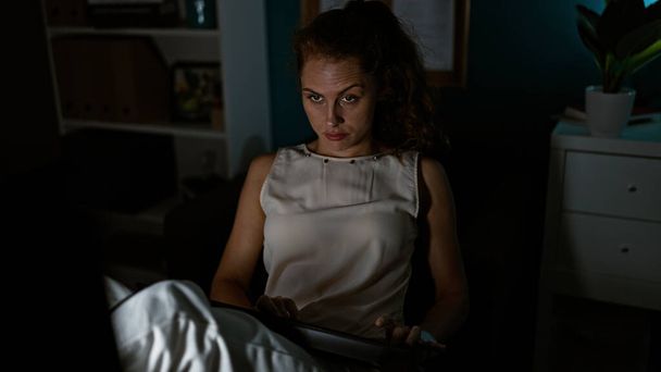Una joven mujer caucásica enfocada en su trabajo nocturno en una oscura oficina iluminada por la luz de su tableta. - Foto, Imagen