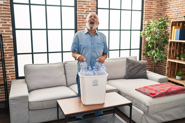 Innenporträt eines wütenden Seniors, frustriert über das Recycling von Plastikflaschen, aggressiv schreiend im heimischen Wohnzimmer - Foto, Bild