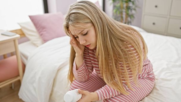 Une jeune femme en détresse examine une bouteille de pilule alors qu'elle est assise sur un lit dans une chambre lumineuse. - Photo, image